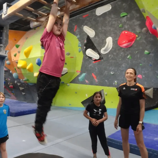 Ein junges Mädchen hängt an einer Holzleiter in einem Fitnessstudio, während zwei Frauen beobachten. Ein Mädchen trägt ein schwarzes Shirt, das andere schwarzes und orangefarbenes. Die Wand hat farbige Formen und hält Klettergriffe.