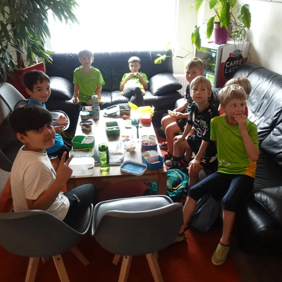 Eine Gruppe von Kindern sitzt um einen Tisch mit Essen, Getränken und einer Box darauf. Einige tragen grüne und weiße T-Shirts. Dahinter steht ein Sofa und Topfpflanzen.
