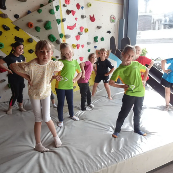 Eine Gruppe von Kindern steht auf einer gepolsterten Oberfläche vor einer Kletterwand mit bunten Handgriffen. Sie tragen Sportbekleidung.