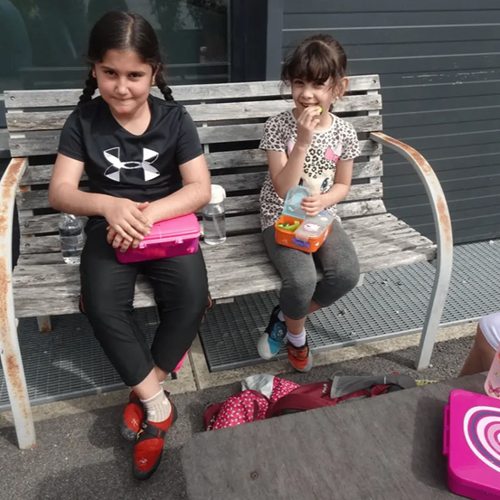 Zwei Mädchen sitzen auf einer Holzbank und essen. Eines hat eine pinke Brotdose, das andere eine orangefarbene. Sie haben Wasserflaschen und Rucksäcke auf der Bank.