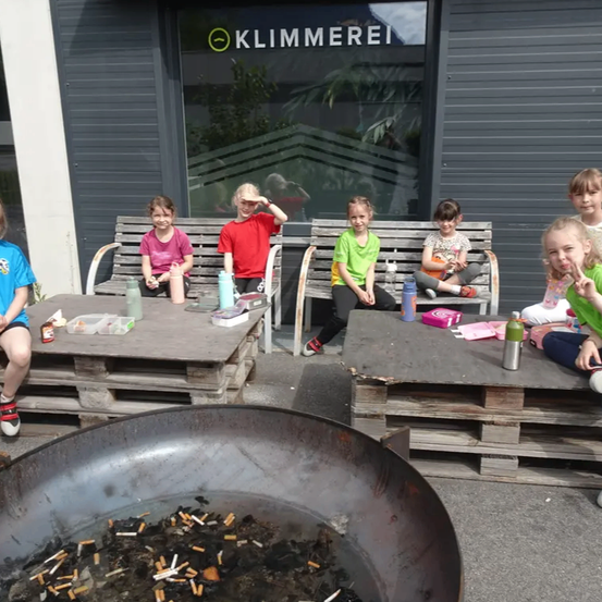 Mehrere Kinder sitzen auf Bänken vor einem Gebäude mit der Aufschrift Klimmerei, umgeben von Gegenständen wie Flaschen und Snacks. Im Vordergrund steht ein großer Wok mit Zigarettenresten.