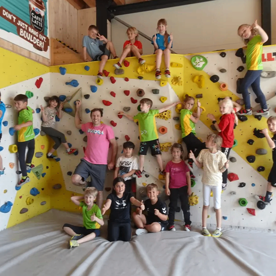 Eine Gruppe von Kindern und Erwachsenen befindet sich in einer Indoor-Kletterhalle. Einige klettern, während andere für ein Foto posieren. Eine Frau in einem rosa T-Shirt steht unter den Kindern.