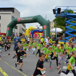 Eine Gruppe von Kindern in grünen Shirts läuft in einem Rennen, gefolgt von mehreren Erwachsenen. Ein grüner Bogen mit dem Wort 'Fohrenburger' steht vorne. Gebäude, Bäume und eine große Menge sind im Hintergrund.
