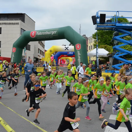Eine Gruppe von Kindern in grünen Shirts läuft in einem Rennen, gefolgt von mehreren Erwachsenen. Ein grüner Bogen mit dem Wort 'Fohrenburger' steht vorne. Gebäude, Bäume und eine große Menge sind im Hintergrund.