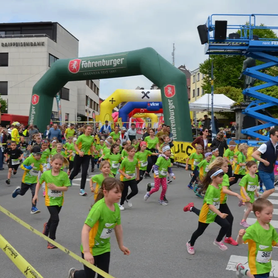 Kinder in grünen Startnummern laufen ein Rennen in der Stadt und passieren einen grünen Torbogen mit der Aufschrift 'Fohrenburger'. Sie werden von Zuschauern angefeuert.