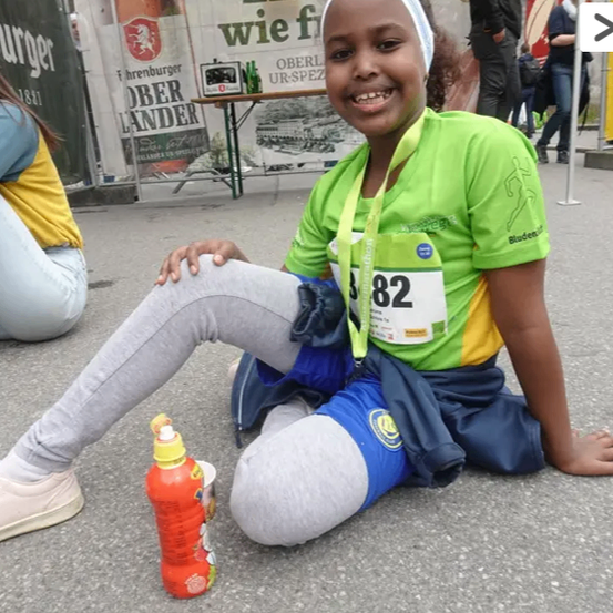 Ein Mädchen mit der Nummer 82 auf dem Shirt sitzt auf dem Boden, lächelt und hält eine Flasche Saft. Im Hintergrund stehen verschwommene Menschen.