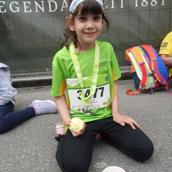 Ein junges Mädchen mit einer Medaille um den Hals sitzt lächelnd auf dem Boden, mit einem Becher Essen vor sich. Ein Rucksack steht neben ihr, und eine Wand mit dem Text 'Legenda' und dem Jahr '1881' ist hinter ihr.