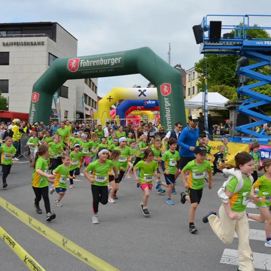 Eine Gruppe von Kindern in grünen und gelben Shirts läuft in einem Marathon. Der Marathon hat einen Bogen mit dem Text 'Fohrenburger' und 'Legendar Seit 1881'. Ein Mann in einem blauen Shirt und einer Kappe führt das Rennen an.