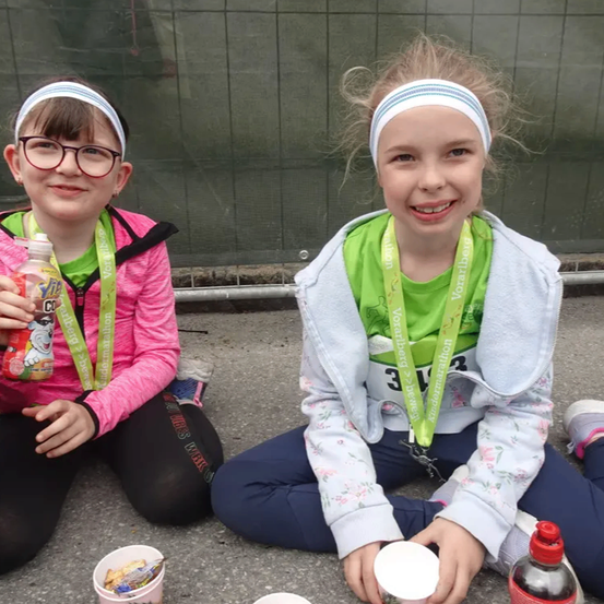 Zwei junge Mädchen sitzen auf dem Boden und lächeln beide. Beide tragen Kopfbänder für Veranstaltungen. Ein Mädchen hält eine Flasche Joghurtgetränk und einen Becher. Das andere Mädchen hat eine Flasche Sportgetränk. Beide sind in Laufausrüstung gekleidet. Hinter ihnen ist ein grüner Zaun zu sehen.