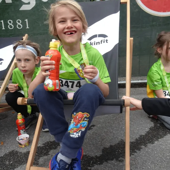 Ein junger Junge mit einer Medaille sitzt auf einem Klappstuhl und hält eine Flasche, während andere in der Nähe sitzen und ein Banner im Hintergrund.