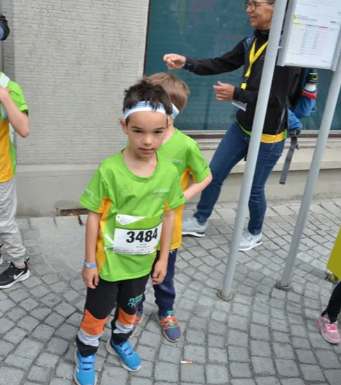 Zwei Kinder in grünen Shirts stehen auf einem gepflasterten Bürgersteig. Eine Frau mit Brille und Rucksack geht hinter ihnen her.