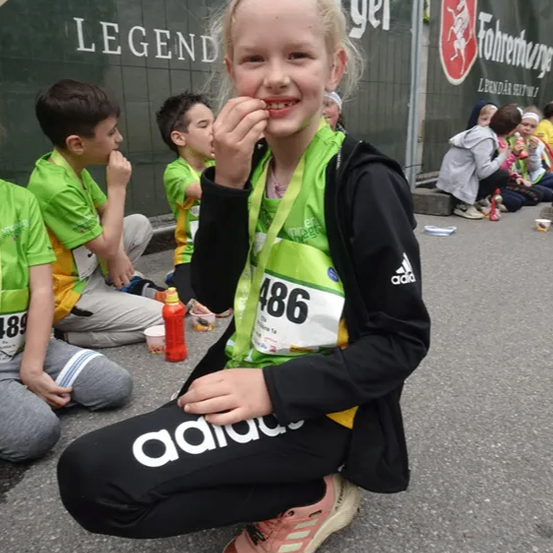 Ein Mädchen mit der Nummer 486 auf dem Shirt sitzt mit einem Lächeln auf dem Boden, während andere Kinder hinter ihr sitzen. Dahinter steht ein Zaun mit dem Wort 'Fohrenburger'.