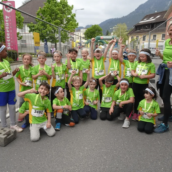 Eine Gruppe junger Athleten in grünen Uniformen posiert für ein Foto, wahrscheinlich nach einem Rennen, mit Nummern auf ihren Trikots und einer lächelnden Frau dahinter.
