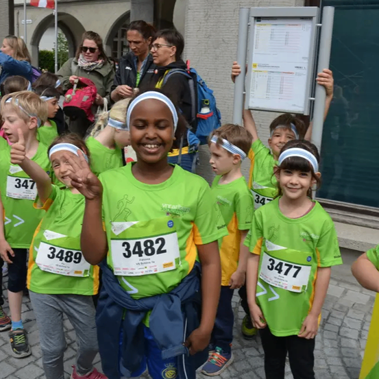 Mehrere Kinder mit grünen T-Shirts und Stirnbändern stehen draußen, manche mit Nummern auf ihren Shirts. Ein Mädchen mit der Nummer 3482 lächelt in die Kamera. Dahinter hält eine Frau ein Schild hoch.