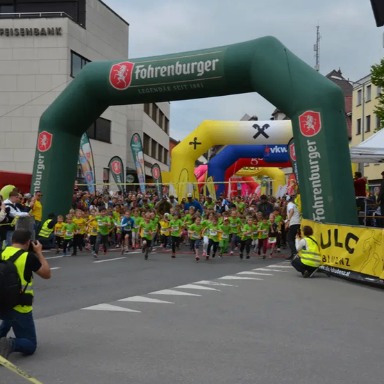 Eine Gruppe von Kindern in grünen Trikots läuft durch einen Bogen bei einem Marathon, mit Zuschauern an der Seite. Ein Fotograf hockt in der Nähe und hält den Moment fest. Hinter den Kindern stehen mehrere Gebäude, von denen eines 'Raiffeisenbank' liest.