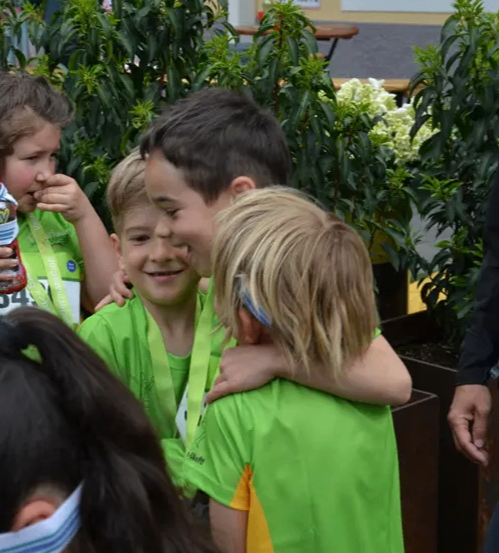 Eine Gruppe von Kindern, einige in grünen Shirts, feiern zusammen mit Medaillen um den Hals. Sie stehen zwischen großen Topfpflanzen.