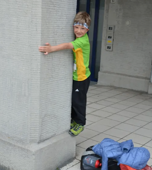Ein junger Junge in einem grün-gelben Shirt, mit einem Stirnband, lehnt sich gegen einen grauen Pfeiler, mit einem Rucksack und einer Jacke auf dem Boden in der Nähe.