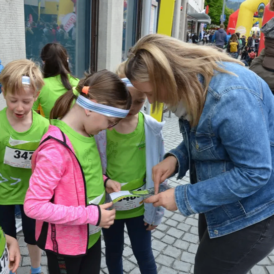 Eine Frau reicht einem Mädchen in einer rosa Jacke ein Papier. Das Mädchen trägt ein grünes Shirt mit der Nummer 347.
