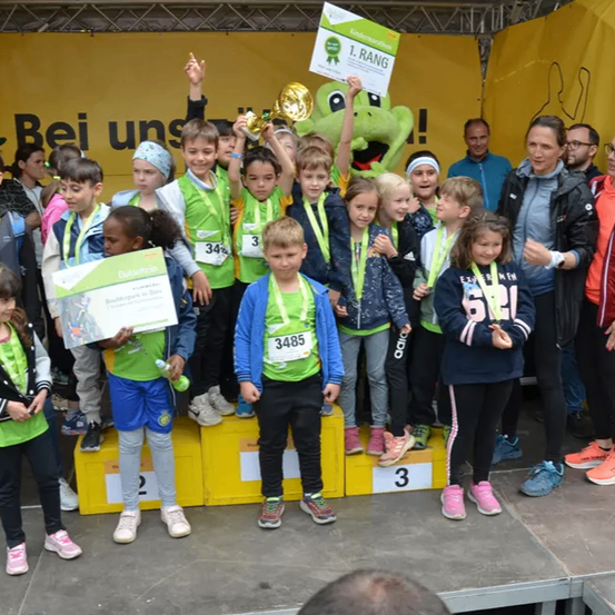 Eine Gruppe von Kindern steht mit Medaillen und Trophäen auf einem Podium und trägt passende grüne Shirts. Dahinter beobachten Erwachsene und andere Kinder, und ein grüner Banner trägt die Aufschrift 'Bei uns'.