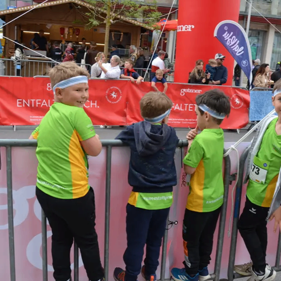 Vier Jungen, die Stirnbänder tragen, stehen hinter einem Zaun, wahrscheinlich bei einem Marathon-Event. Ein Junge trägt ein grün-gelbes Shirt, ein anderer eine blaue Jacke. Zuschauer stehen hinter ihnen.