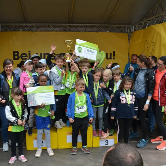 Mehrere Kinder stehen mit ihren Trophäen auf dem Podest und lächeln für die Kamera. Ein großer gelber Banner mit dem Text 'Bei uns' im Hintergrund.