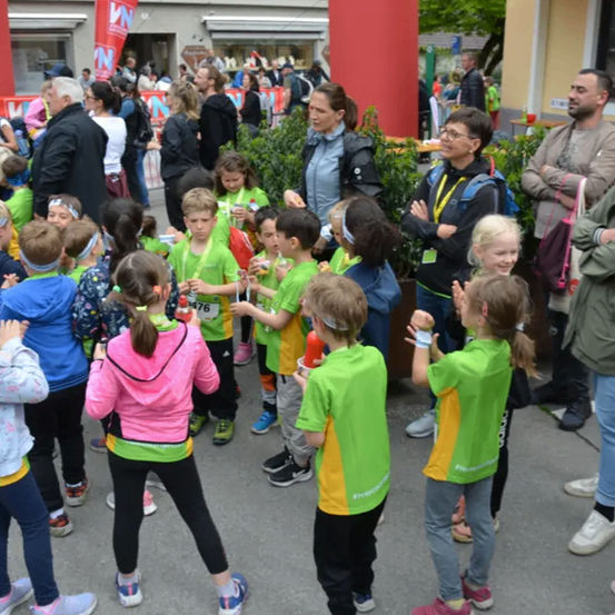 Eine Gruppe von Kindern in grünen und gelben Shirts steht im Kreis, einige klatschen und andere halten Getränke. Sie sind von Erwachsenen umgeben, einige beobachten und andere führen Gespräche. Der Hintergrund zeigt eine geschäftige Outdoor-Veranstaltung mit Bannern und einer großen Menschenmenge.