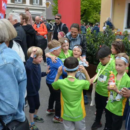 Eine Gruppe von Kindern in grünen Shirts steht in einem Kreis, trägt Stirnbänder und hält eine Flasche. Erwachsene und andere Kinder sind um sie herum versammelt. Im Hintergrund befinden sich Gebäude und Grünpflanzen.