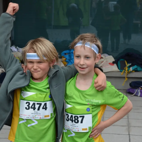 Zwei junge Jungen in grünen T-Shirts mit den Nummern 3474 und 3487 stehen zusammen und posieren für ein Foto, mit einem verschwommenen Hintergrund eines Gebäudes und Personen.