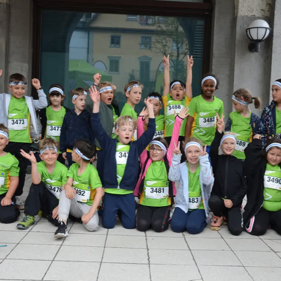 Eine Gruppe von Kindern in grünen T-Shirts mit Nummern posiert für ein Foto vor einem Gebäude. Sie lächeln und machen verschiedene Handgesten. Das Gebäude hat eine Glastür und Fenster.