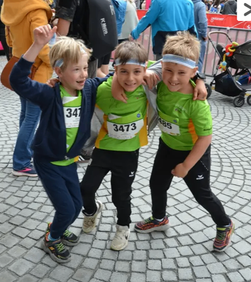 Drei Kinder in grünen Shirts mit den Nummern 347, 3473 und 985 stehen zusammen auf einer Kopfsteinpflasterstraße. Dahinter sind Erwachsene, darunter einer mit einem Rucksack.