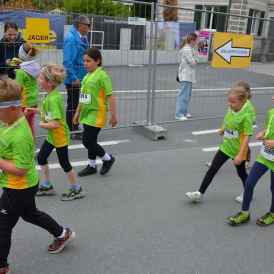 Mehrere Kinder nehmen an einem Rennen teil, tragen grüne und gelbe Trikots mit Nummern. Sie laufen auf einer Straße, mit einem Zaun und einer Frau dahinter stehend.
