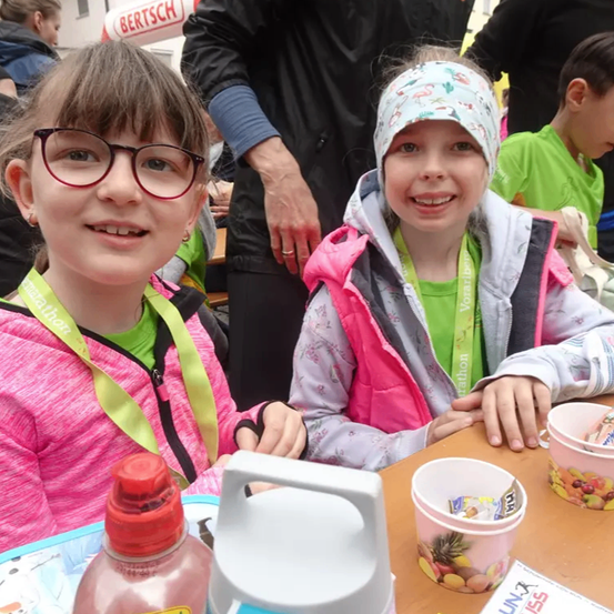 Zwei Mädchen sitzen an einem Tisch. Sie lächeln und halten Becher mit Saft. Sie tragen Ausweiskarten. Hinter ihnen stehen andere Leute.