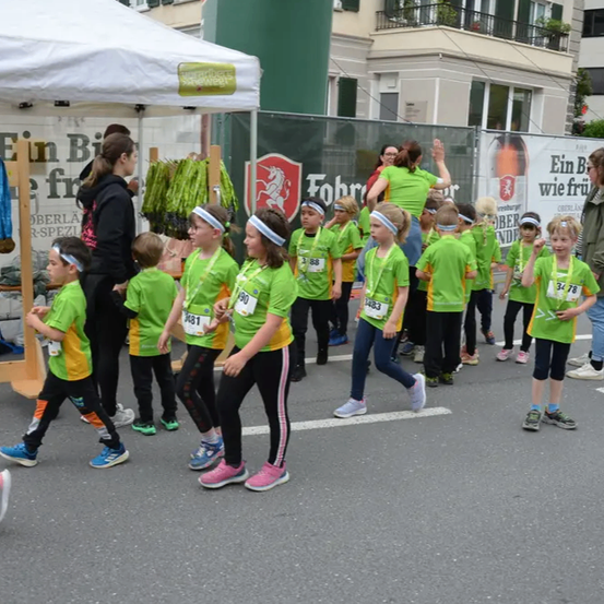 Kinder in grünen und gelben Uniformen mit Nummern gehen in einer Reihe auf einer Straße. Ein Zelt mit einem Banner steht dahinter, und eine erwachsene Frau steht in der Nähe. Ein Banner mit einem Logo ist am Zaun. Gebäude sind im Hintergrund.