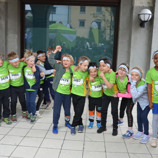 Eine Gruppe von Kindern, die passende grüne Hemden und Stirnbänder mit Nummern tragen, posiert für ein Foto vor einem Gebäude. Sie lächeln und scheinen an einem Rennen teilzunehmen.