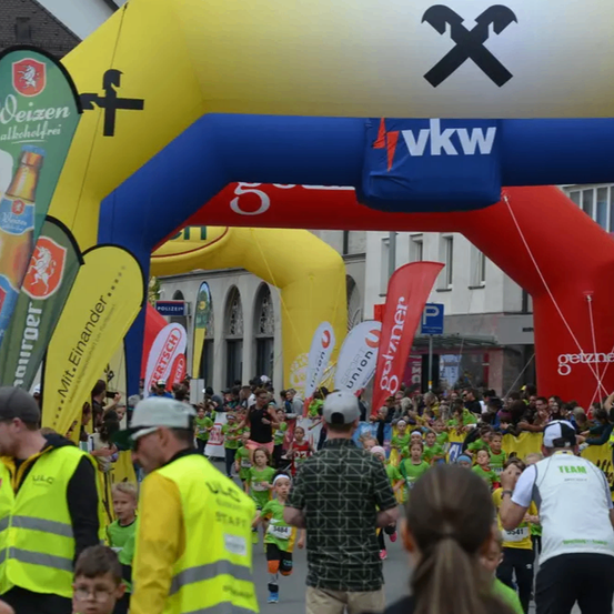 Marathonteilnehmer laufen durch einen dekorierten Bogen mit Flaggen und Sponsoren, einschließlich eines für Weizen-Bier. Die Menge und Offizielle tragen neonfarbene Westen.