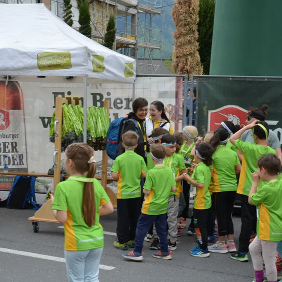 Eine Gruppe von Kindern in grünen T-Shirts steht um ein Zelt herum, auf dem eine Flasche Fahrenbruger ausgestellt ist. Eine Frau steht hinter einem Tisch mit einem Rucksack. Einige Leute stehen hinter ihnen.