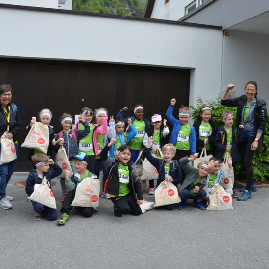 Eine Gruppe von Kindern und Erwachsenen, die grüne Shirts tragen, steht draußen, wahrscheinlich in einem Park, und hält Taschen. Sie posieren für ein Foto, wobei einige von ihnen die Hände heben. Hinter ihnen befindet sich ein Gebäude mit einer Garage und ein Fahrrad, das an der Seite geparkt ist.
