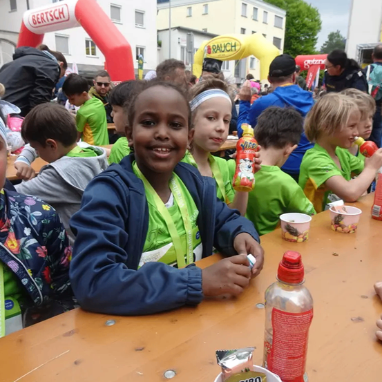 Eine Gruppe von Kindern in grünen Shirts sitzt an einem Tisch mit Getränken und Essen. Ein Junge lächelt in der Mitte. Ein Banner hinter ihnen lautet Rauch.