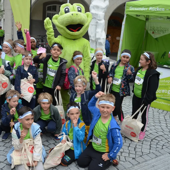 Eine Gruppe von Kindern in grünen Shirts posiert für ein Foto mit einem Frosch-Maskottchen bei einer Veranstaltung.