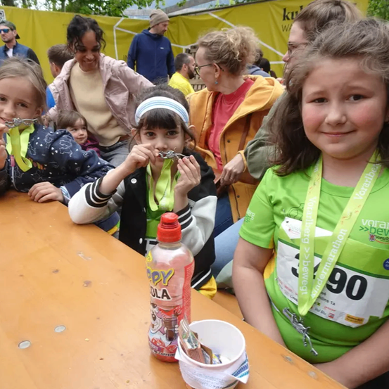 Eine Gruppe von Kindern mit Medaillen und grünen Nummernbändern sitzt lächelnd am Tisch. Eine Getränkeflasche und ein Becher stehen auf dem Tisch. Dahinter schaut eine Menge Menschen zu, mit einem Banner im Hintergrund.