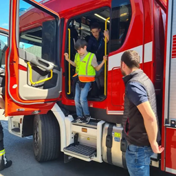 Ein Junge in einer Warnweste steigt mit Hilfe eines Mannes auf einen roten Feuerwehrwagen, unter einem klaren Himmel.