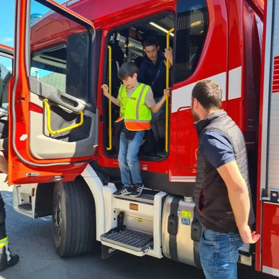 Ein Junge in einer Warnweste steigt mit Hilfe eines Mannes auf einen roten Feuerwehrwagen, unter einem klaren Himmel.