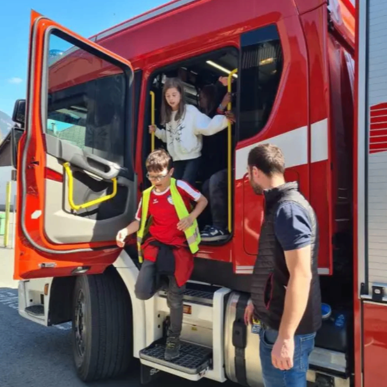 Ein Kind in einer Warnweste steigt mit Hilfe eines Erwachsenen die Stufen eines Feuerwehrwagens hinauf. Eine Frau in einem weißen Shirt hilft von innen aus dem Wagen.