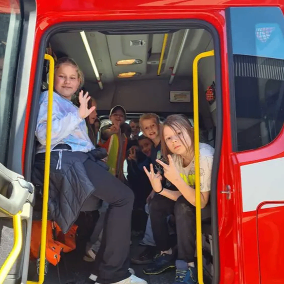 Mehrere Kinder sitzen in einem roten Bus. Ein Mädchen vorne lächelt und macht ein Friedenszeichen. Ein Mädchen neben ihr macht ein Foto mit ihrem Handy. Der Bus hat gelbe Handläufe und Sitze im Inneren.
