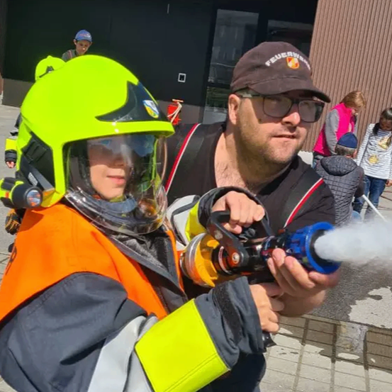 Ein Junge in einer Feuerwehruniform hält einen Wassersprüher mit herausspritzendem Wasser. Er steht neben einem Mann mit einer Kappe und einer Brille. Hinter ihnen sind weitere Personen anwesend.