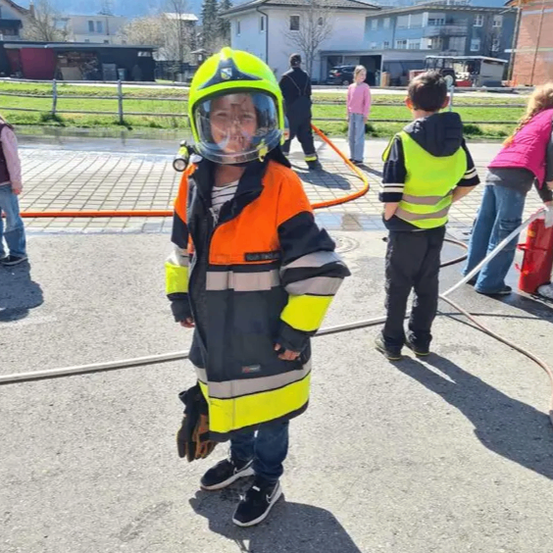 Ein Kind in einer Feuerwehruniform steht auf der Straße mit Bergen im Hintergrund. Andere Kinder in ähnlichen Uniformen sind in der Nähe, und Erwachsene sind anwesend, einschließlich einer Person mit einem Feuerlöscher.