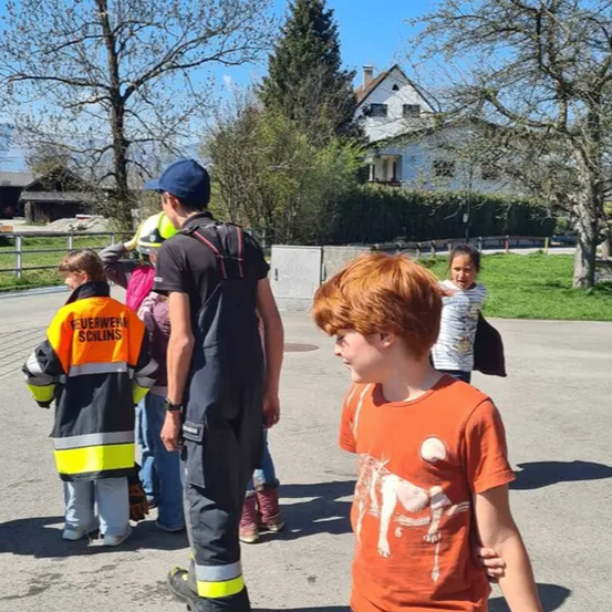 Eine Gruppe von Kindern und Erwachsenen steht im Freien. Ein Junge in einem orangefarbenen T-Shirt spricht mit einem Feuerwehrmann.