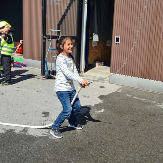 Ein junges Mädchen sprüht Wasser aus einem Schlauch vor einem Gebäude, in der Nähe einer Person in einer gelben Weste.