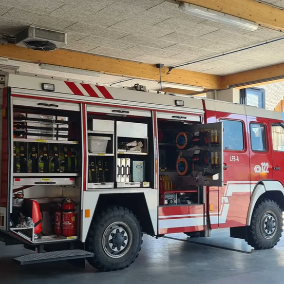 Ein roter und weißer Feuerwehrwagen steht in einer Garage mit geöffneten Türen und zeigt verschiedene Ausrüstungsgegenstände und Werkzeuge.