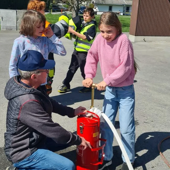 Ein Mann hilft einem Mädchen, einen Feuerlöscher zu bedienen, während mehrere Kinder zusehen. Sie befinden sich auf einem Parkplatz mit Gebäuden und einem Baum im Hintergrund.